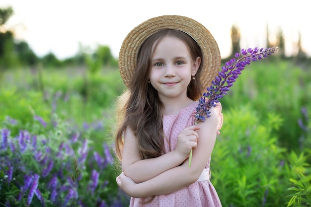 Happy little girl in straw hat holds lupine flower in her hands at sunset Summer vacation