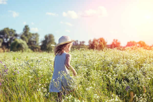 Happy little girl in straw hat walking in the flowers field on a sunny summer day