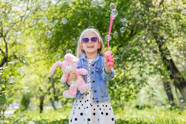 A happy little girl in summer with sunglasses inflates soap bubbles holds a toy in her hands