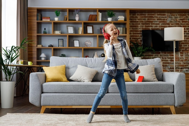 Happy little girl wearing red headphones dancing and singing jumping on a floor Cute child enjoy listening favorite music tracks in mobile application Home leisure happy childhood