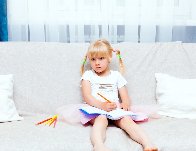 Happy little girl with blond hair draws in a notebook at home. Preschool girl is engaged in drawing. The concept of a happy childhood.