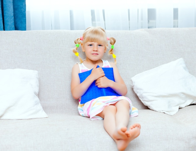 Photo happy little girl with blond hair is reading a book sitting on a sofa in the living room of her house. distance learning.