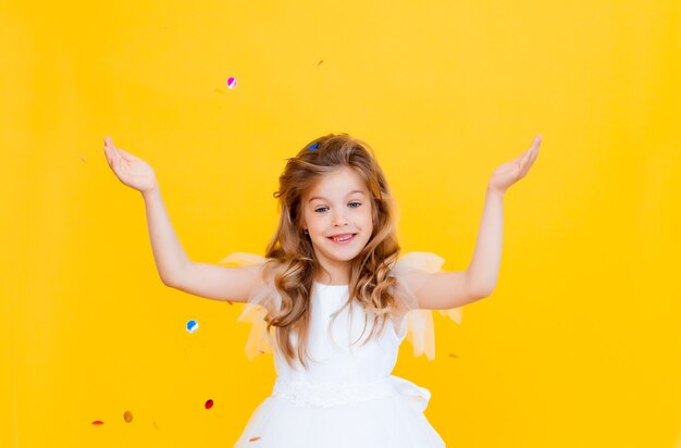 Happy little girl with blond hair and in a white dress catches confetti on a yellow background, holiday concept