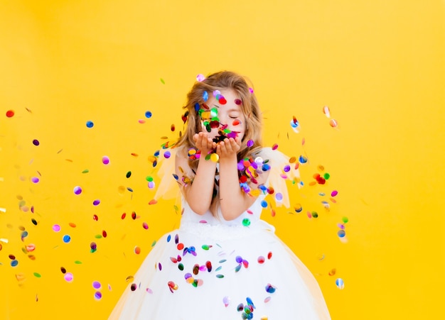 Happy little girl with blond hair and in a white dress catches confetti on a yellow background, holiday concept