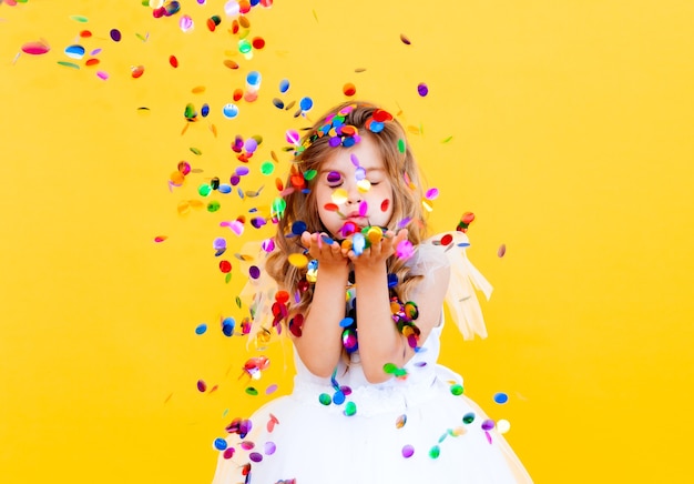 Happy little girl with blond hair and in a white dress catches confetti on a yellow background, holiday concept