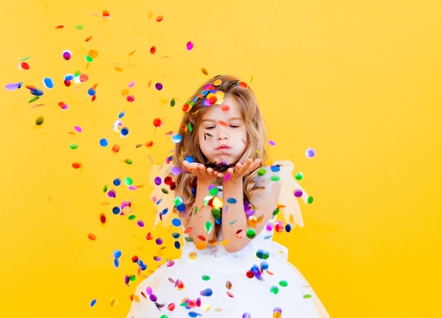 Happy little girl with blond hair and in a white dress catches confetti on a yellow background, holiday concept