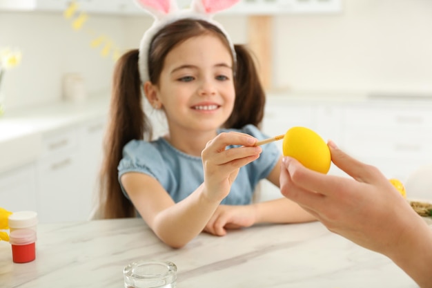 Happy little girl with bunny ears headband and her mother painting Easter egg in kitchen