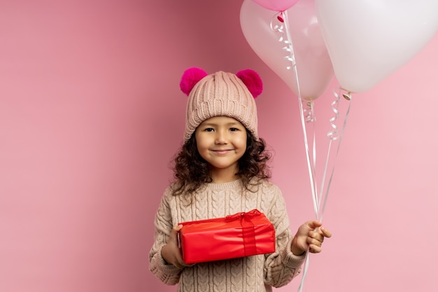 Happy little girl with curly hair wearing warm sweater, winter hat with fluffy pompoms, holding red gift box and balloons, smiling isolated