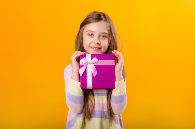 Happy little girl with long hair holding a pink gift box on a yellow space isolate