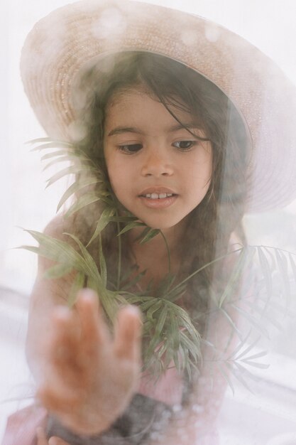 Happy little girl with a potted indoor flower in her hands looks out of the window from the house