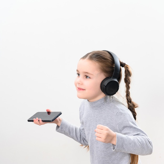 Happy little girl with wireless headphones and phone portrait of girl with pigtails on white background with headphones and smartphone