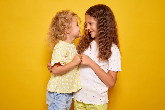 Photo happy little girls with cheerful toothy smiles playing together indoors holding hands embracing looking at each other with positive emotions standing isolated over yellow background