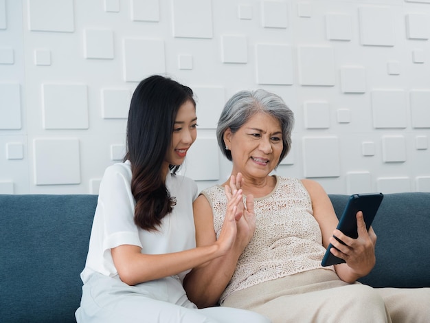 Happy lovely Asian senior woman mother white hair and beautiful young female daughter waving greeting at digital tablet screen while sit on couch in white room Meet family by video call at home