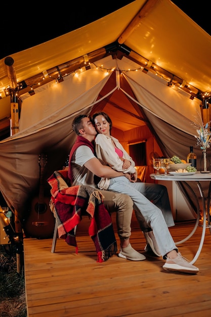 Happy lovely couple relaxing in glamping on summer evening and drinking wine near cozy bonfire Luxury camping tent for outdoor recreation and recreation Lifestyle concept