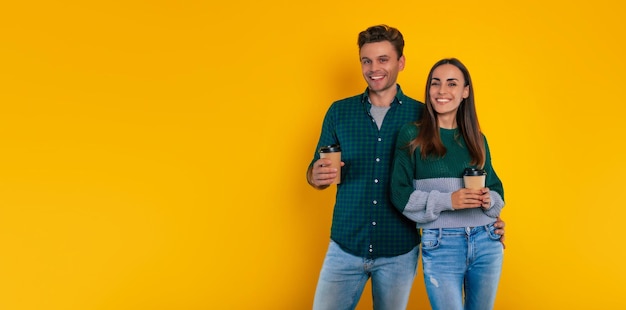 Happy lovely young couple with cups of coffee or tea in hands wearing casual clothes, hugging each other and laughing together while they isolated on yellow background