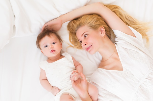 Happy loving family concept. Beautiful mother playing with her baby girl in the bedroom. They smiling and hugging together on white bed linens, top view