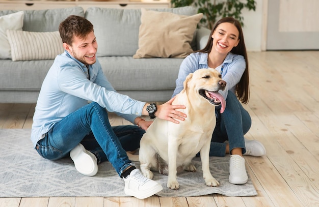 Happy Loving Family Concept. Portrait of cheerful spouses sitting on the floor in modern apartment, spending time together with dog. Man and woman owners playing with labrador pet at home