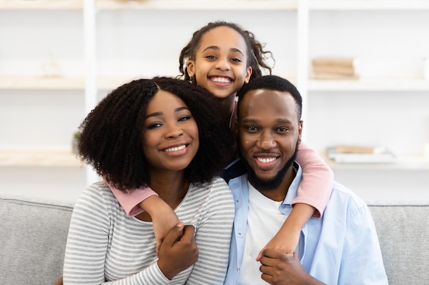 Happy Loving Family Concept. Portrait of excited African American girl hugging her mum and daddy from back, parents sitting on sofa in living room, posing for photo looking at camera at home