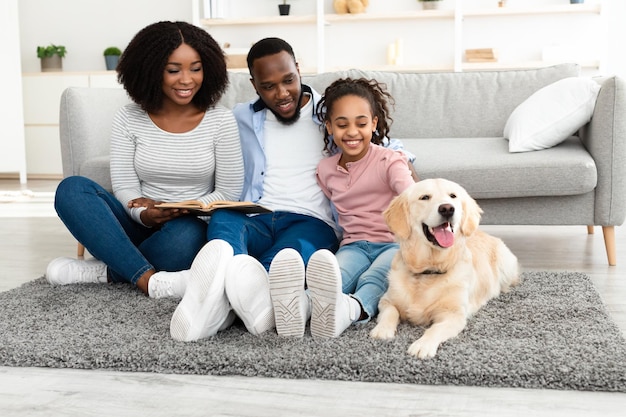 Happy Loving Family. Portrait of cheerful black mum, dad and daughter reading paper book or holding photo album togetherm sitting on the rug floor carpet with domestic dog, girl patting labrador