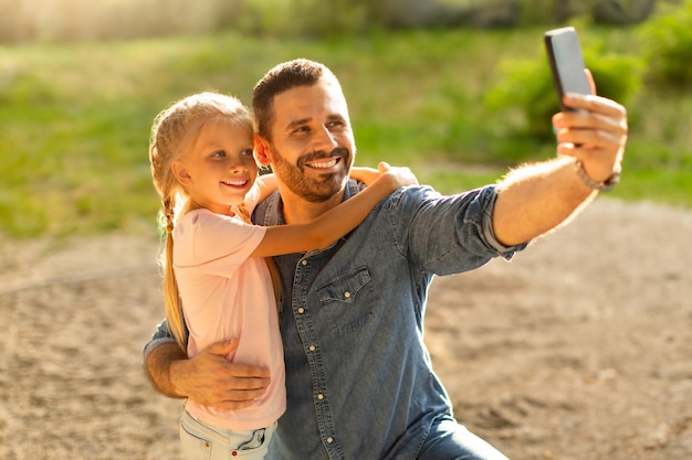 Happy loving father taking selfie with his pretty daughter while walking in park at family day