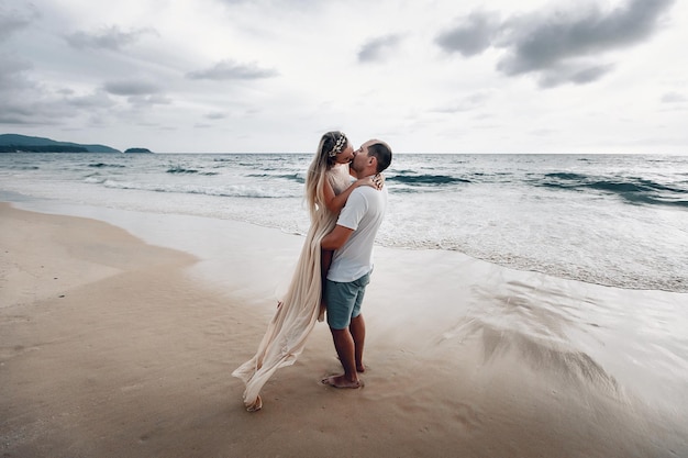 Happy loving husband and wife dressed in white kissing on an exotic beach, the man hugs the girl as he lifts her from the ground.