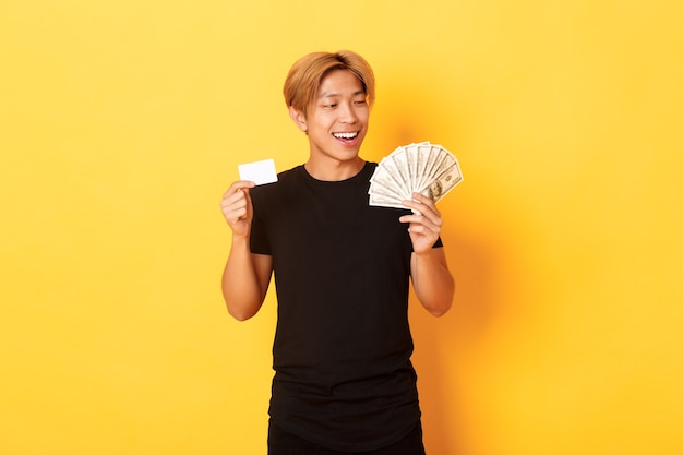 Happy and lucky asian guy looking pleased at money while showing credit card and cash, standing yellow wall