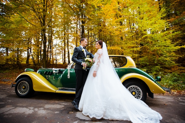 Happy luxury wedding couple kissing and embracing near retro with bouquet car in autumn