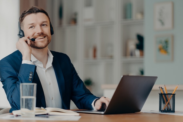 Happy male entrepreneur in suit working from home, successful businessman in headset talking with colleagues online through web conference while sitting at his cozy workplace. Remote work at home