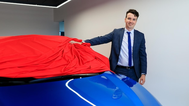 A happy male manager during the preparation of the car before the sale The car is under a red cloth