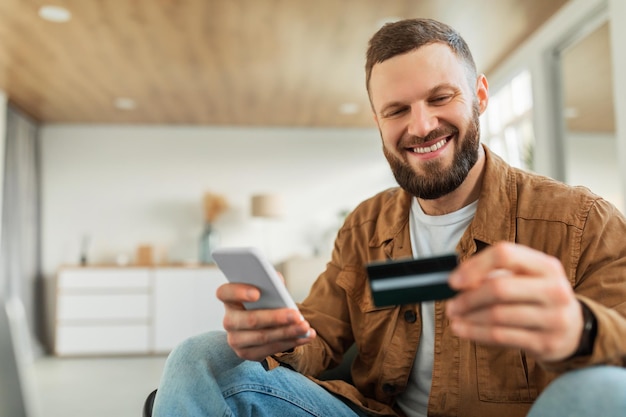 Happy male shopping using smartphone and credit card sitting indoors