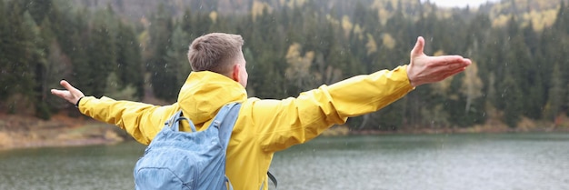 Happy male tourist with open arms stands on shore of lake and looks at mountains outdoors