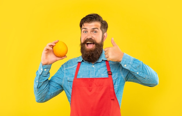 Happy man in apron giving thumb to fresh orange citrus fruit yellow background fruiterer