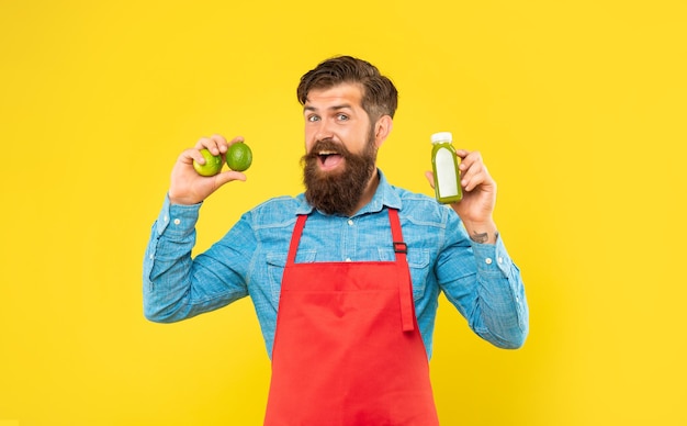 Happy man in apron holding limes and juice bottle yellow background, juice barman.