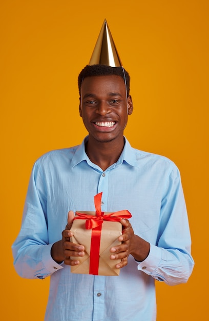 Happy man in cap holding gift box with red ribbons, yellow background. Smiling male person got a surprise, event or birthday celebration