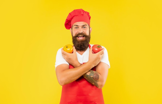 Happy man in cooking apron and toque holding tomatoes in crossed hands yellow background cook