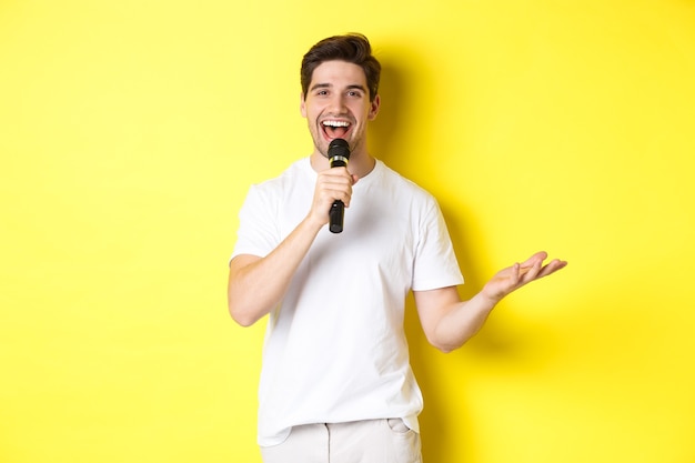 Happy man entertainer performing, talking in microphone, making speech or stand-up show, standing over yellow wall