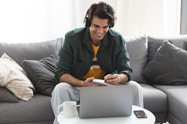 Happy man entrepreneur in headphones speaking looking at laptop and taking notes in notepad. Young male student or coach talking by video conference sitting at home office. Online learning concept