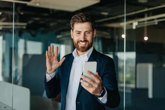 Happy man entrepreneur working in modern office talking on cellphone having video chat and waving hand at webcam