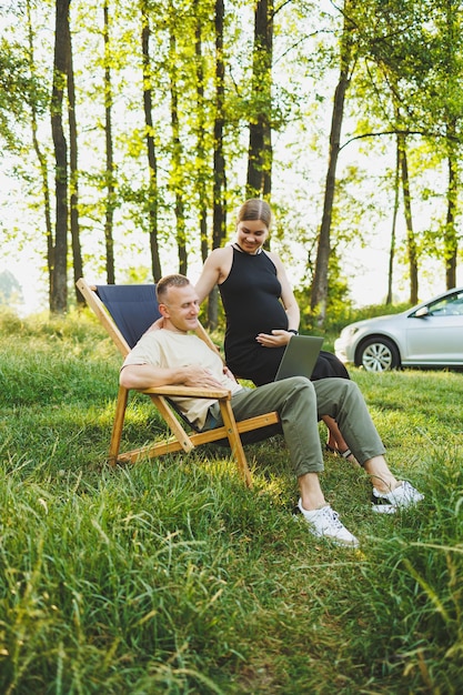 A happy man and his pregnant wife are working on a laptop while sitting in nature A young couple is relaxing in nature and watching a movie on a laptop