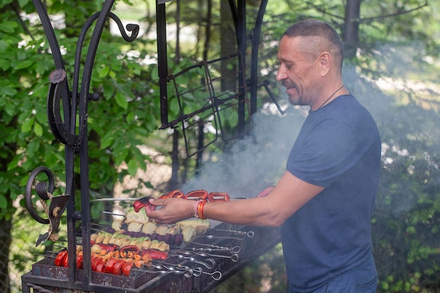 Happy man is cooking vegetable kebab on skewers on the grill in the yard