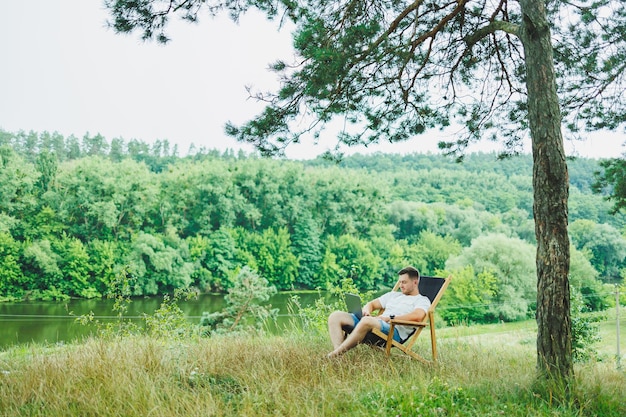 A happy man is relaxing in nature and working remotely on a portable laptop Remote work while on vacation Working on a laptop while sitting in a chair in nature