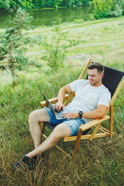 A happy man is relaxing in nature and working remotely on a portable laptop Remote work while on vacation Working on a laptop while sitting in a chair in nature