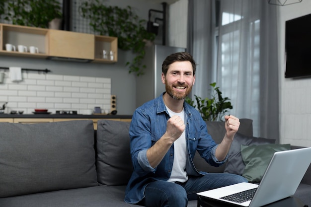 Happy man looks at the camera and smiles rejoices in victory and success works at home on a laptop