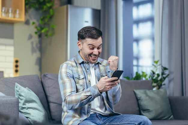 The happy man looks at the phone at home and rejoices in success and victory