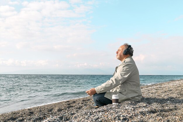 Photo happy man sitting in lotus pose on beach meditating and listening music or podcast