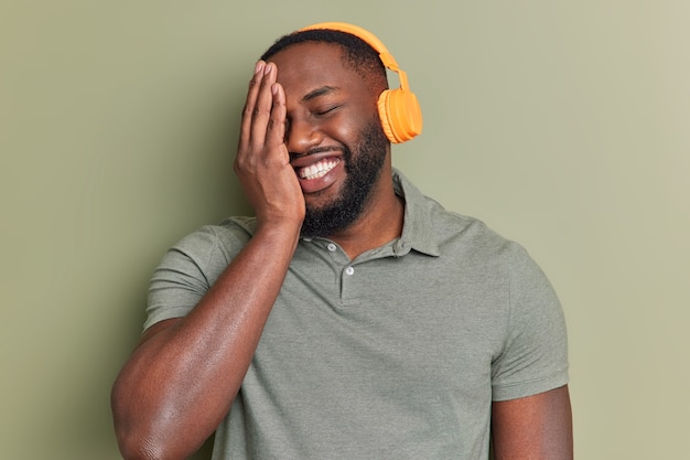 Happy man smiles broadly makes face palm has cheerful look listens music via orange wireless headphones enjoys good sound quality dressed in basic t shirt poses indoor