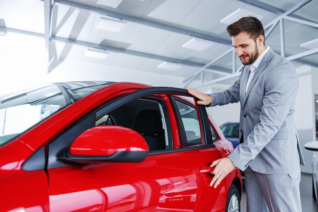 Happy man standing next to a brand new car in car salon and opening door.