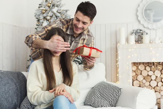 Happy man surprising girl with Christmas gift, closing her eyes from back, having fun on winter holiday, copy space