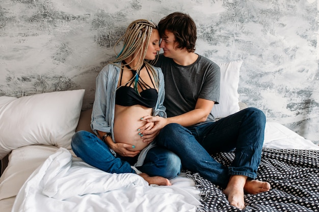 Happy man touching belly of smiling pregnant woman lying on bed at home