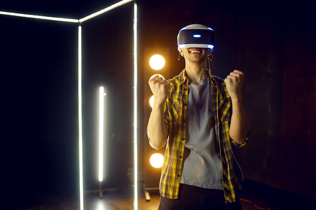 Photo happy man using virtual reality headset and gamepad in luminous cube, front view. dark playing club interior, spotlight on background, vr technology with 3d vision